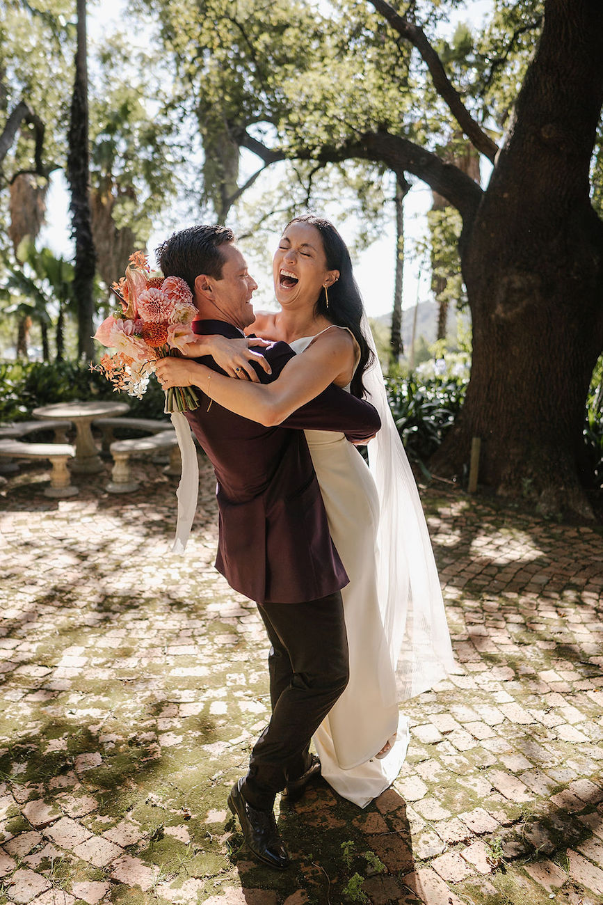 A newlywed couple embraces and smiles joyfully outdoors, with the bride holding a bouquet and wearing a white dress and veil. Sunlight filters through trees in the background.