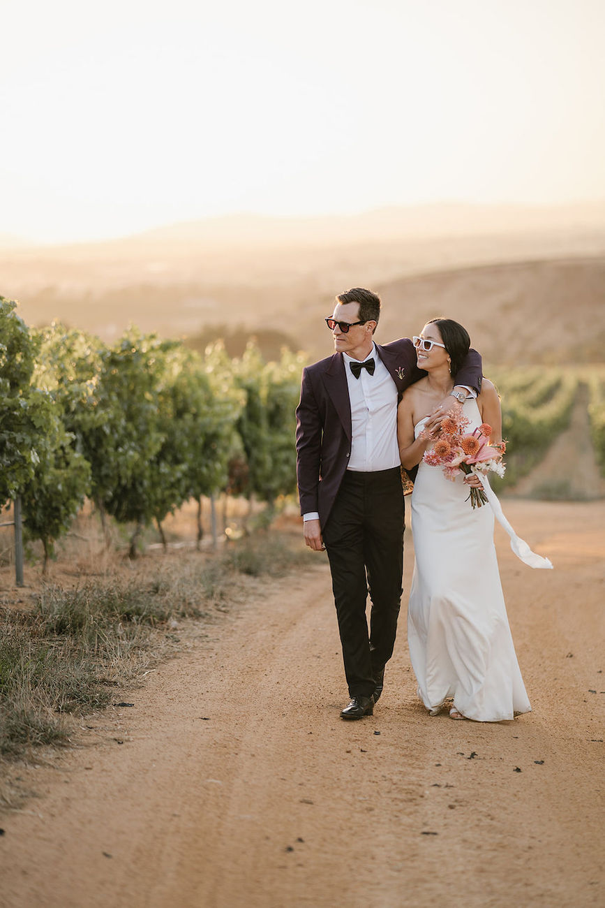 A bride and groom walk arm in arm down a dirt path through a vineyard, dressed in formal wedding attire and holding a bouquet.