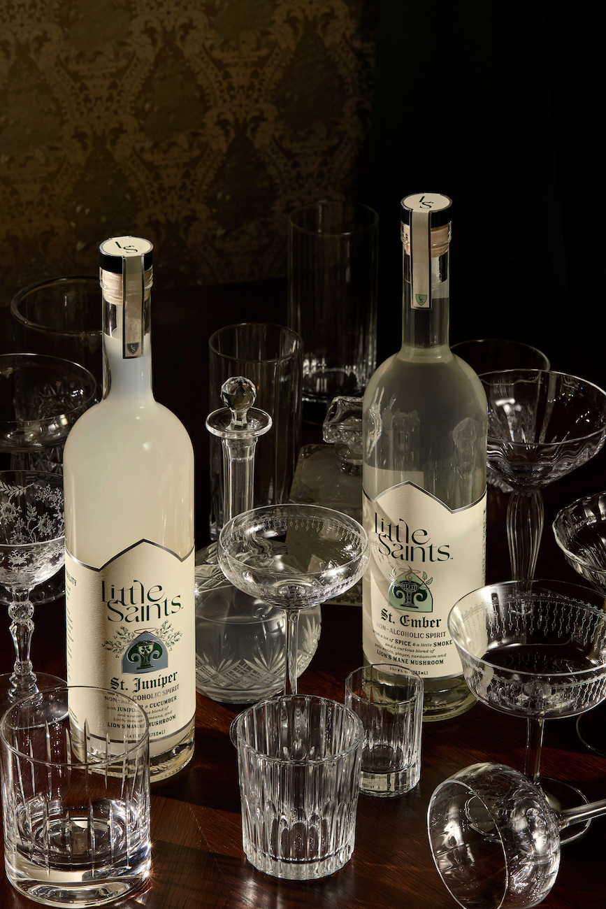 Two bottles of Tutis artis liqueur are displayed on a wooden surface surrounded by various empty crystal glasses and decanters.