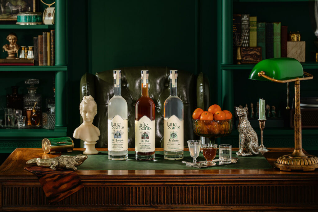 Four bottles of liquor on a wooden desk with glasses, a green desk lamp, books, a bust, a silver dog figurine, and a bowl of oranges in a richly decorated study.