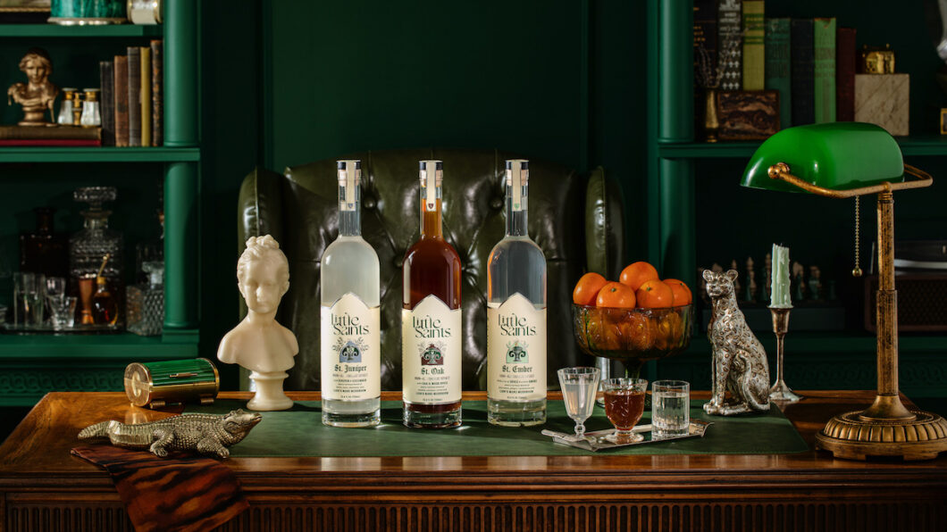 Four bottles of liquor on a wooden desk with glasses, a green desk lamp, books, a bust, a silver dog figurine, and a bowl of oranges in a richly decorated study.