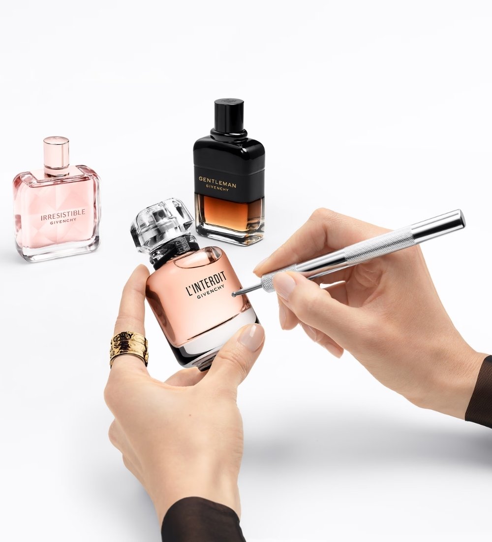 A person holds a Givenchy perfume bottle and writes on it with a silver pen, with two other perfume bottles in the background.