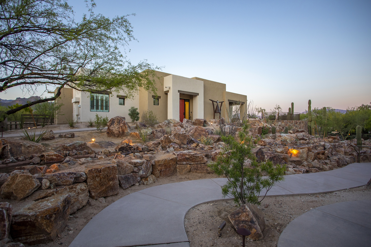 A modern, single-story house with a light brown exterior sits amid a desert landscape with rocks, cacti, and a winding concrete path at dusk.