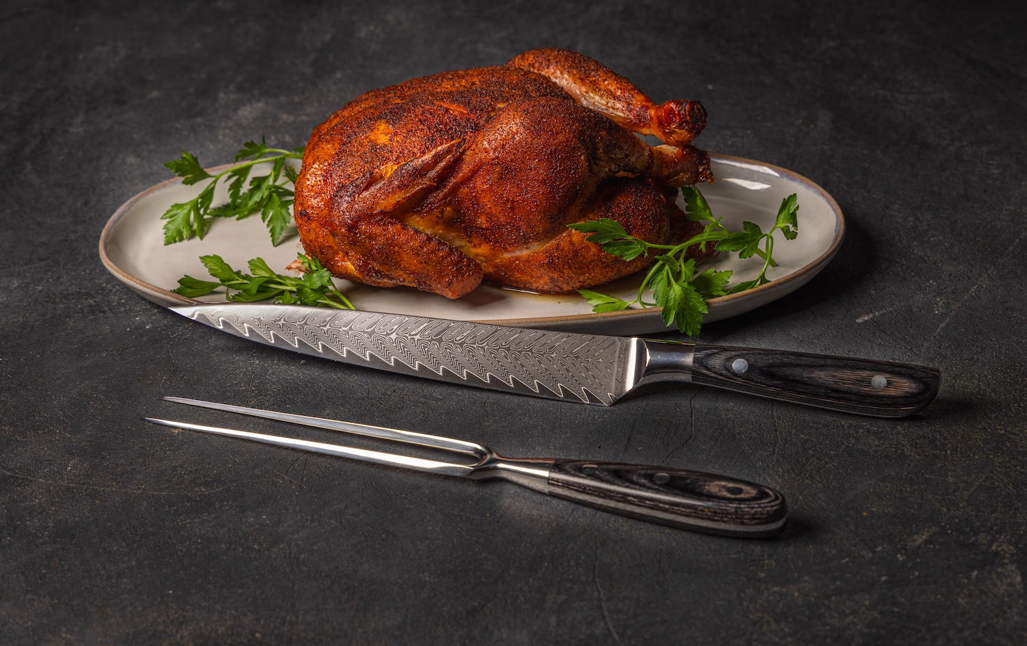 A roasted chicken on an oval plate garnished with parsley, with a carving knife and fork placed in front on a dark surface.
