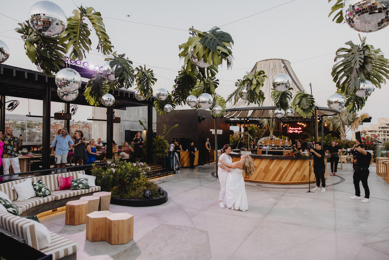 A couple dances in the center of an outdoor venue decorated with hanging plants and disco balls, while guests watch and relax on lounge seating.
