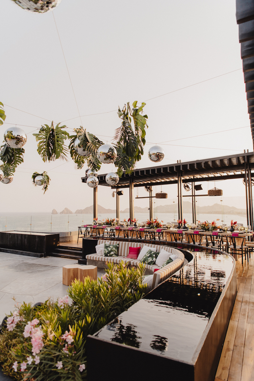 Outdoor rooftop venue with wooden flooring, long dining tables set for an event, plants, and hanging mirrored spheres and greenery, overlooking a hazy seascape.