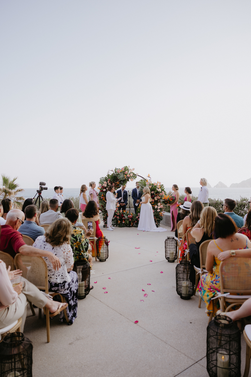 Outdoor wedding ceremony with guests seated, a couple and officiant standing under a floral arch, and flower petals scattered on the ground.