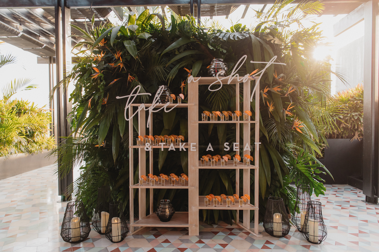 A wooden shelf with small orange drinks is set against lush greenery, with neon signs reading "take a shot" and "take a seat." Lanterns are placed on the floor nearby.