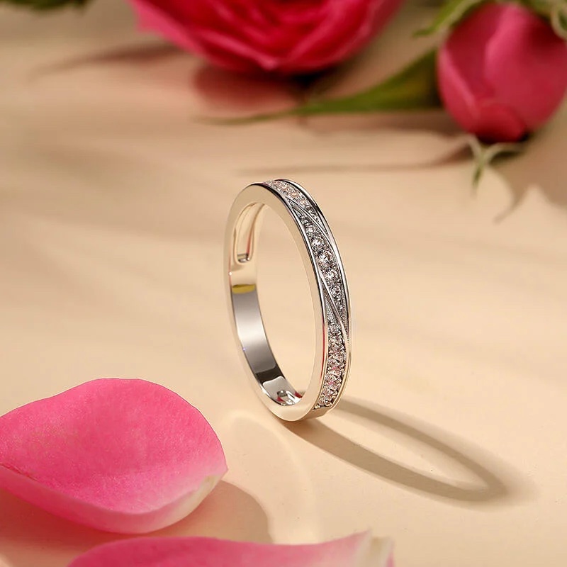 A silver ring with embedded small diamonds is displayed upright on a beige surface, surrounded by pink rose petals and flowers.