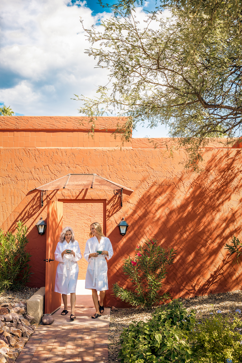 Two people in white robes exit a red-orange building under a wooden awning, holding cups, with plants and a tree nearby under a partly cloudy sky.