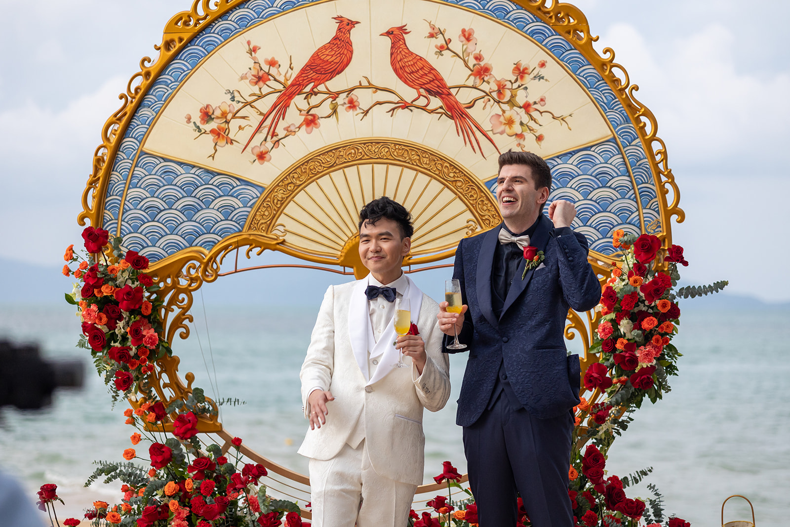 Two men in suits stand smiling with drinks in hand in front of a decorative floral and bird backdrop on a beach.