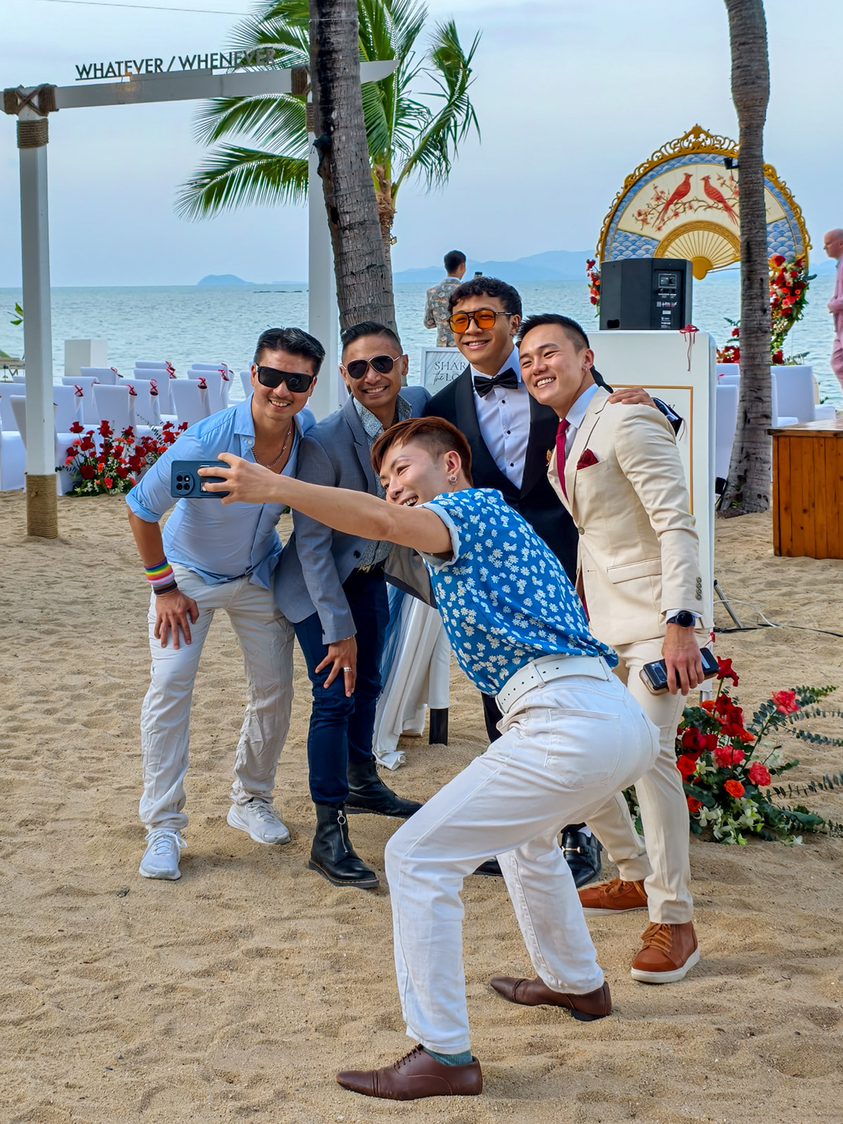 Five men dressed in formal and semi-formal attire take a group selfie on a sandy beach with wedding decorations and the sea in the background.
