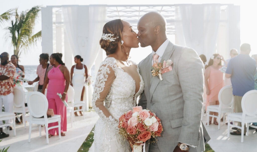 A bride and groom kiss at their wedding ceremony, with the bride holding a bouquet of pink roses and guests seated in the background.