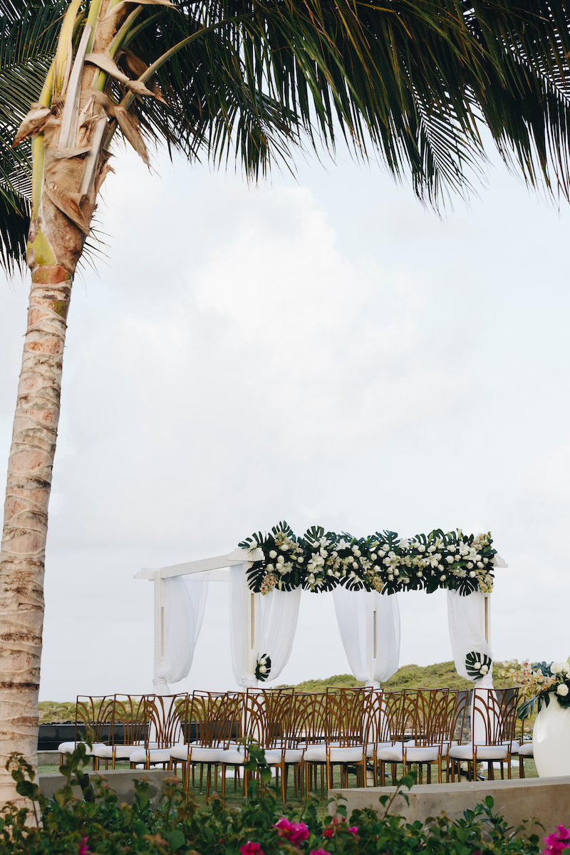A decorated outdoor wedding altar with white drapes and floral arrangements is set up near a palm tree, with rows of empty chairs facing it.