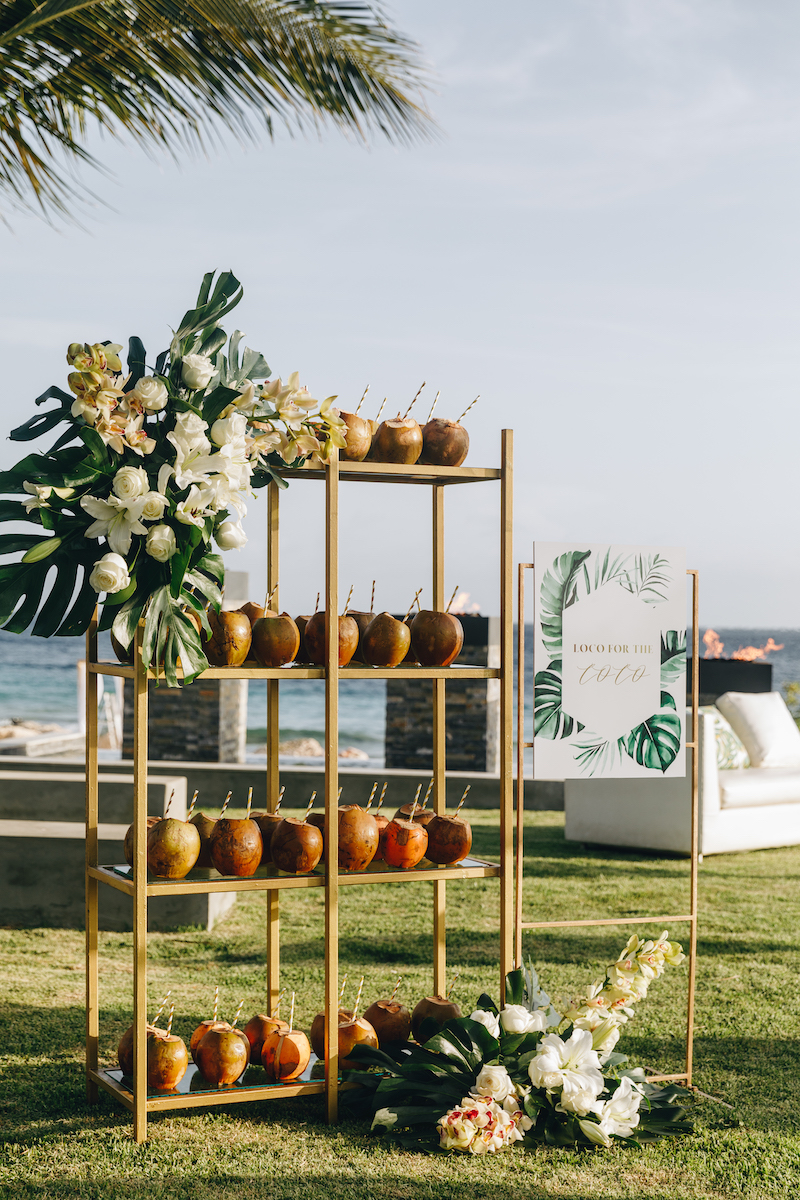 A bamboo shelf displays coconuts with straws and floral arrangements, set on grass near the ocean with a decorative sign beside it.
