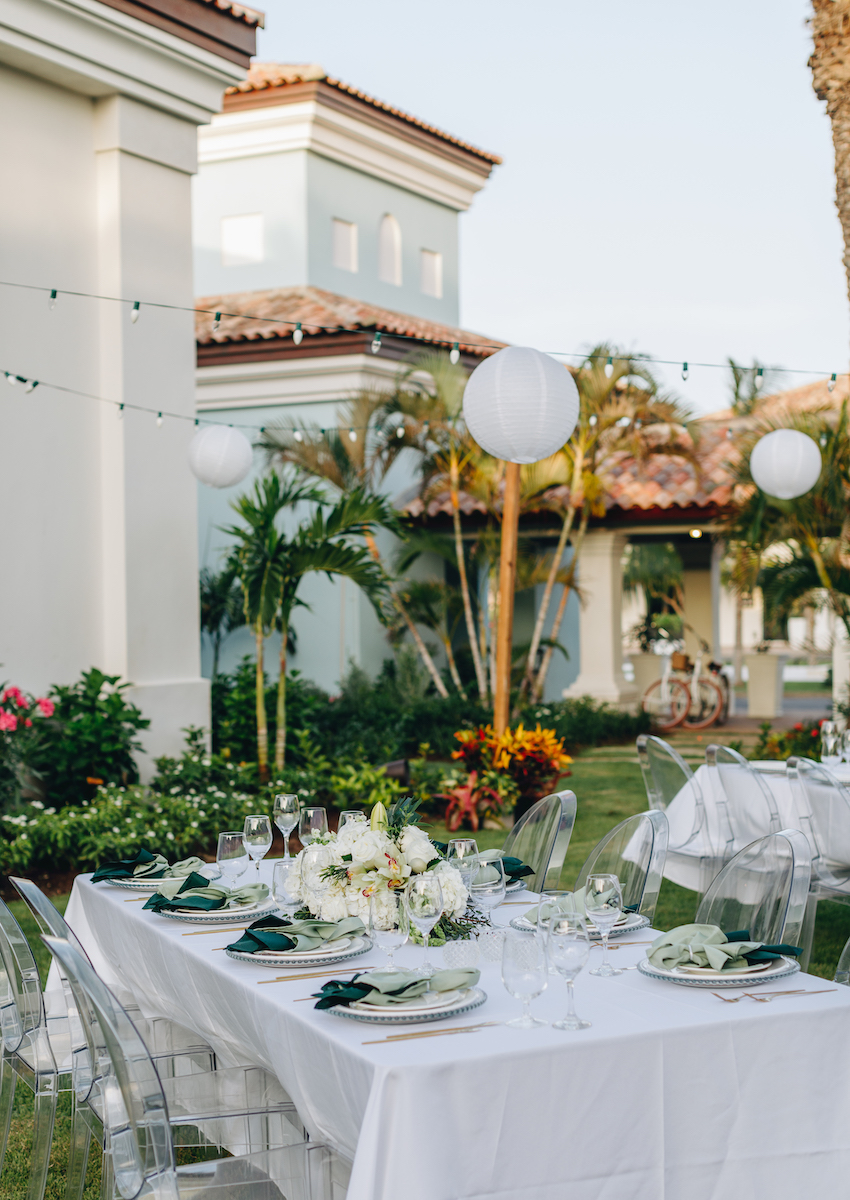 Outdoor table set for a formal event with white tablecloth, floral centerpiece, and place settings, surrounded by greenery and string lights, near a Mediterranean-style building.