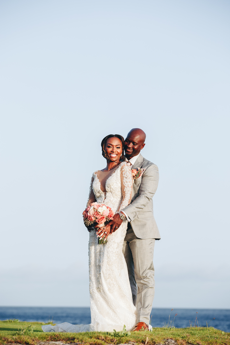 A bride and groom stand outdoors, smiling and embracing, with the ocean and sky in the background. The bride holds a bouquet of flowers.