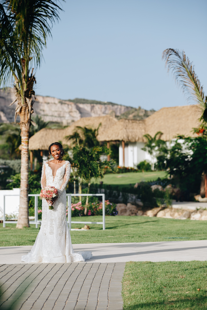 A bride in a white dress holding a bouquet stands outdoors on a sunny day, with palm trees and thatched-roof buildings in the background.