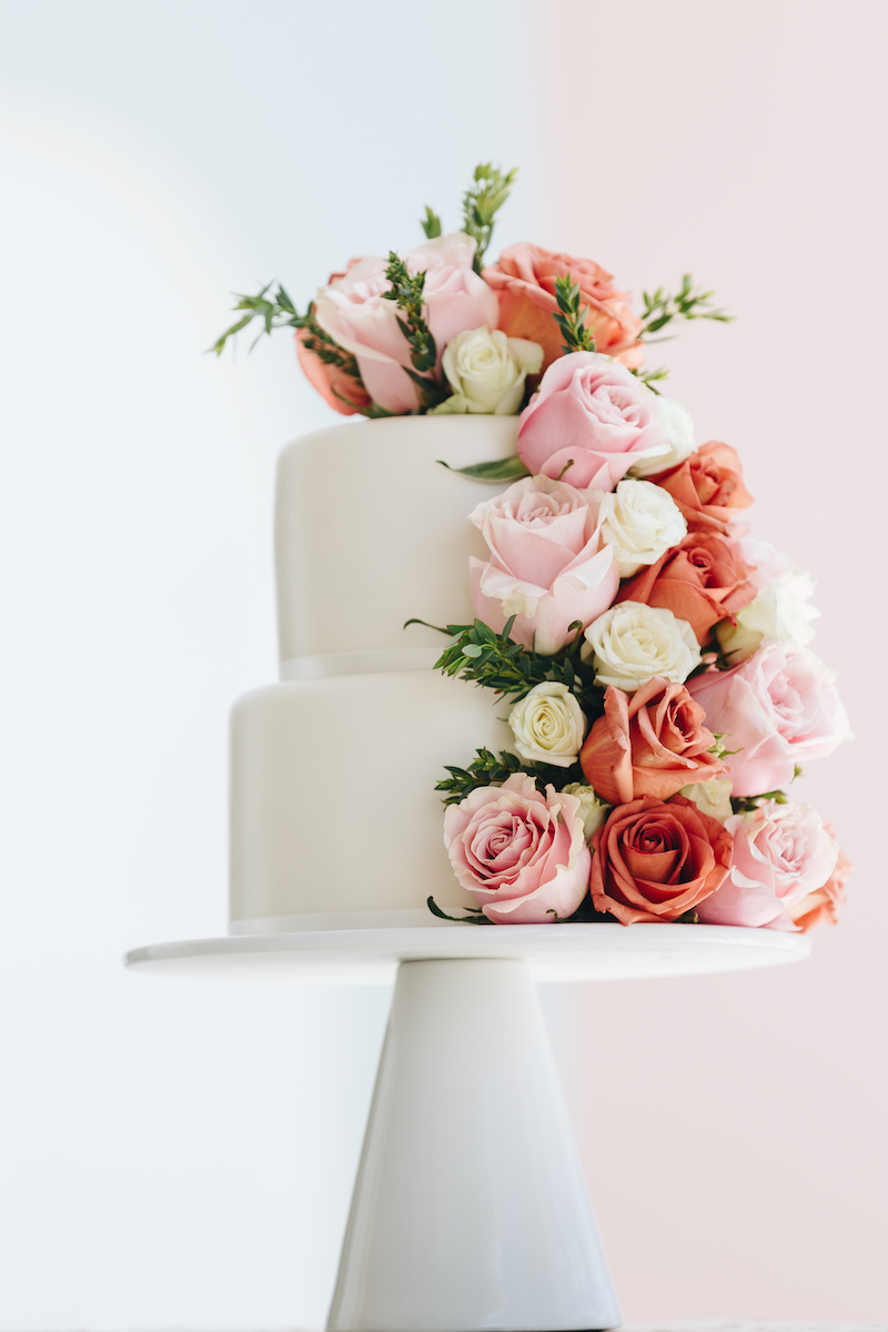 Two-tiered white cake decorated with cascading pink, peach, and white roses, displayed on a white cake stand against a soft pastel background.