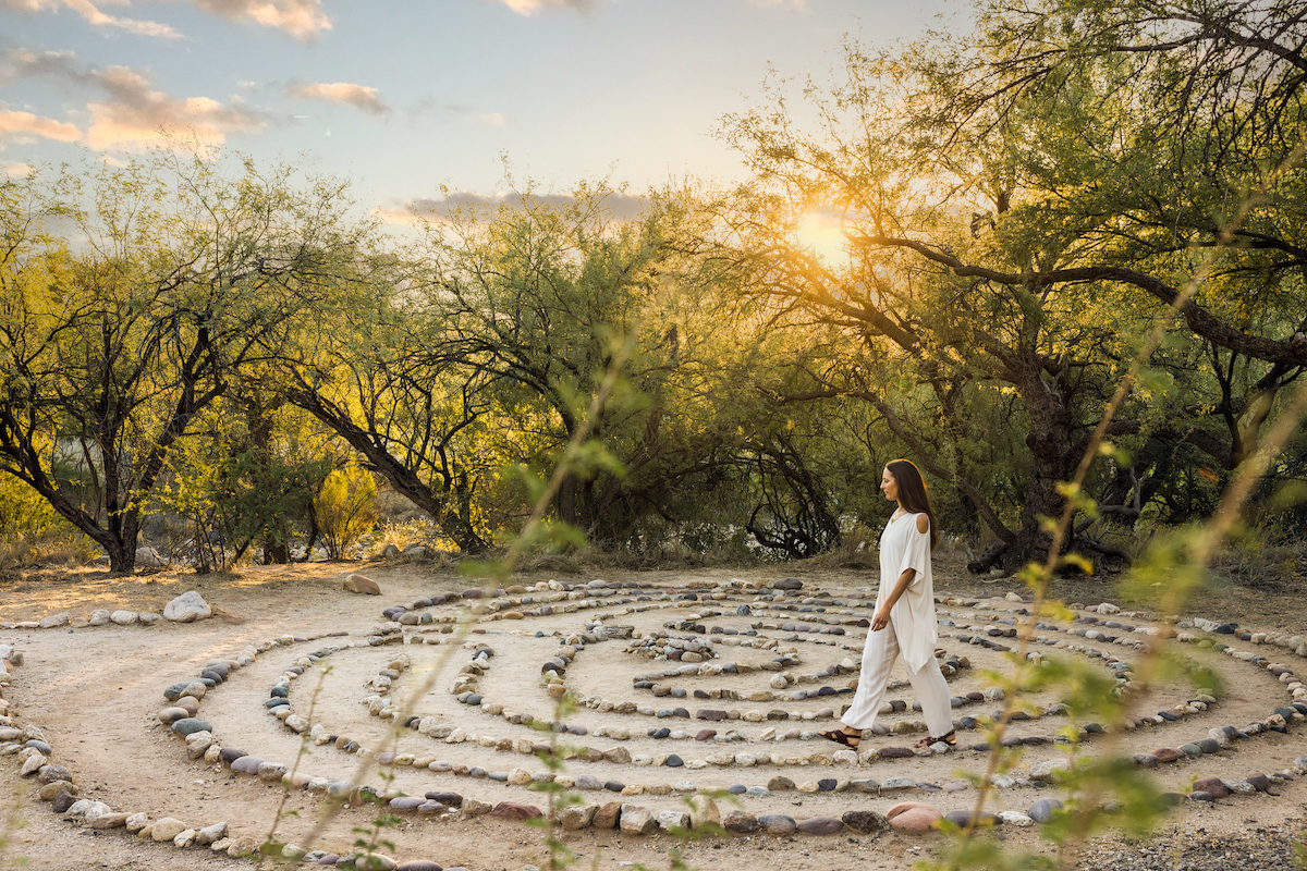 A person in white clothing walks through a stone labyrinth outdoors at sunset, surrounded by trees and natural vegetation.
