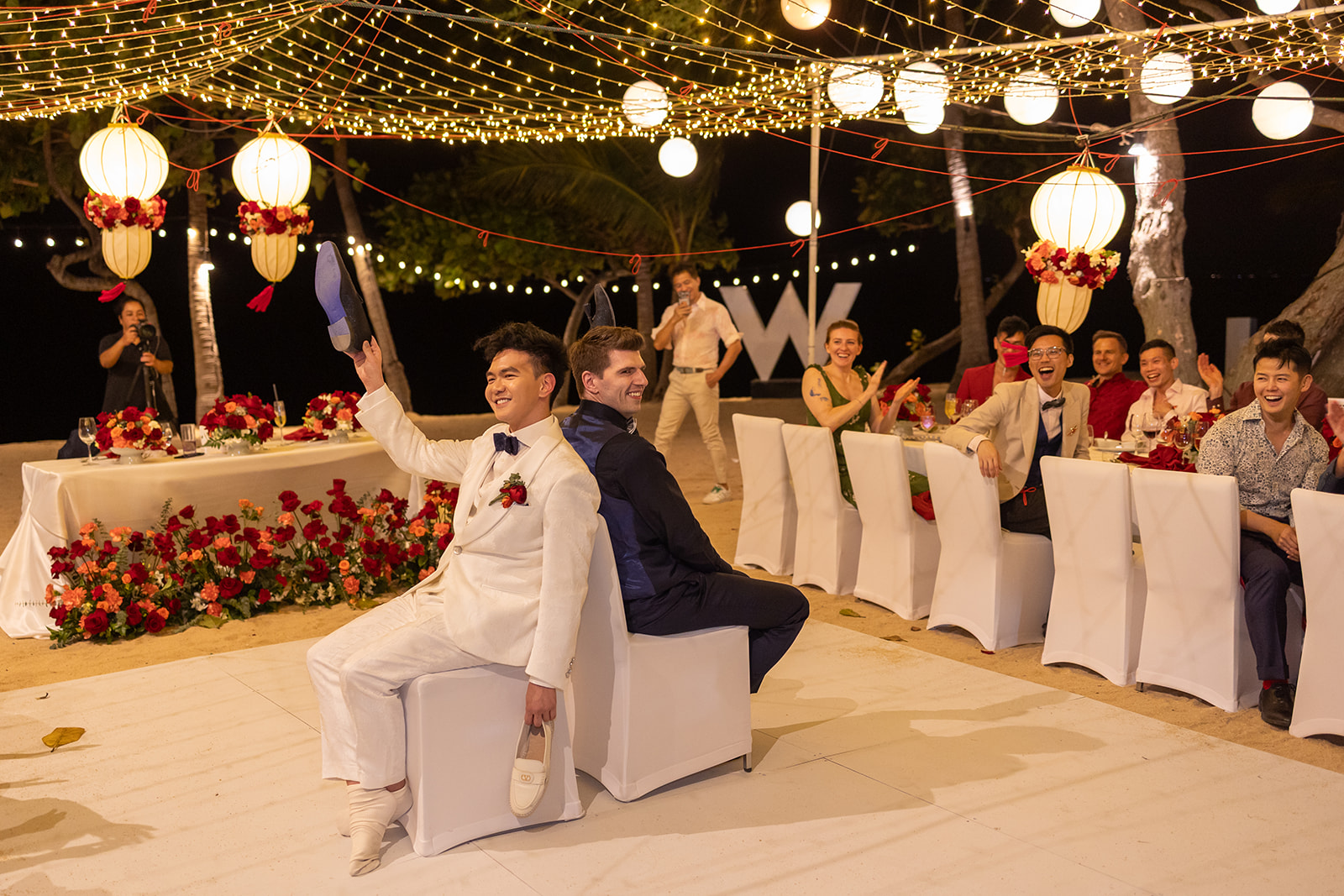 Two grooms sit back-to-back during their wedding reception, each holding up a shoe, surrounded by guests under string lights and lanterns.