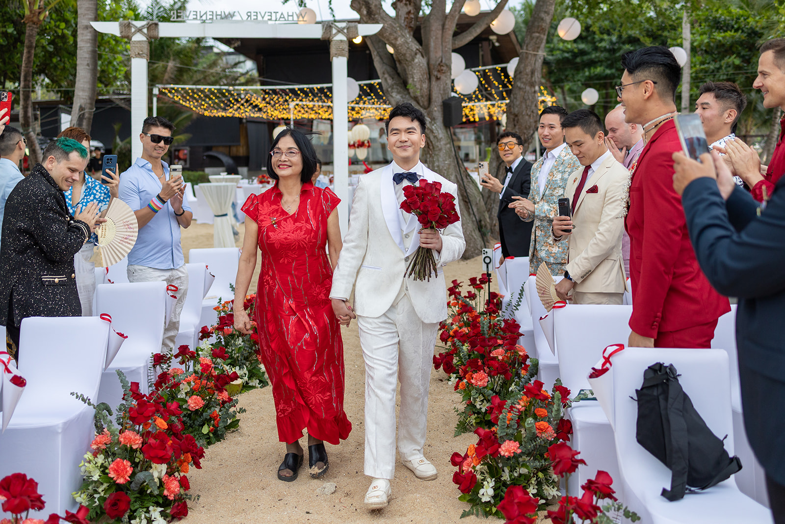 A man in a white suit holding red roses walks down an outdoor aisle with a woman in a red dress, surrounded by guests seated on white chairs decorated with red flowers.
