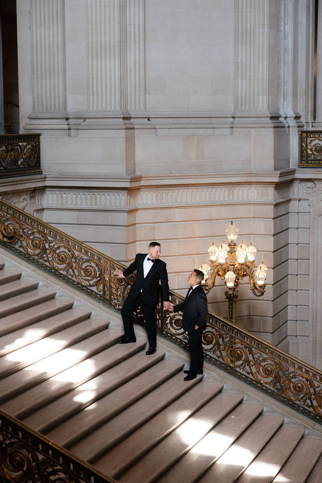 Two men in formal wear stand on an ornate staircase inside a grand building with large columns and decorative lighting.