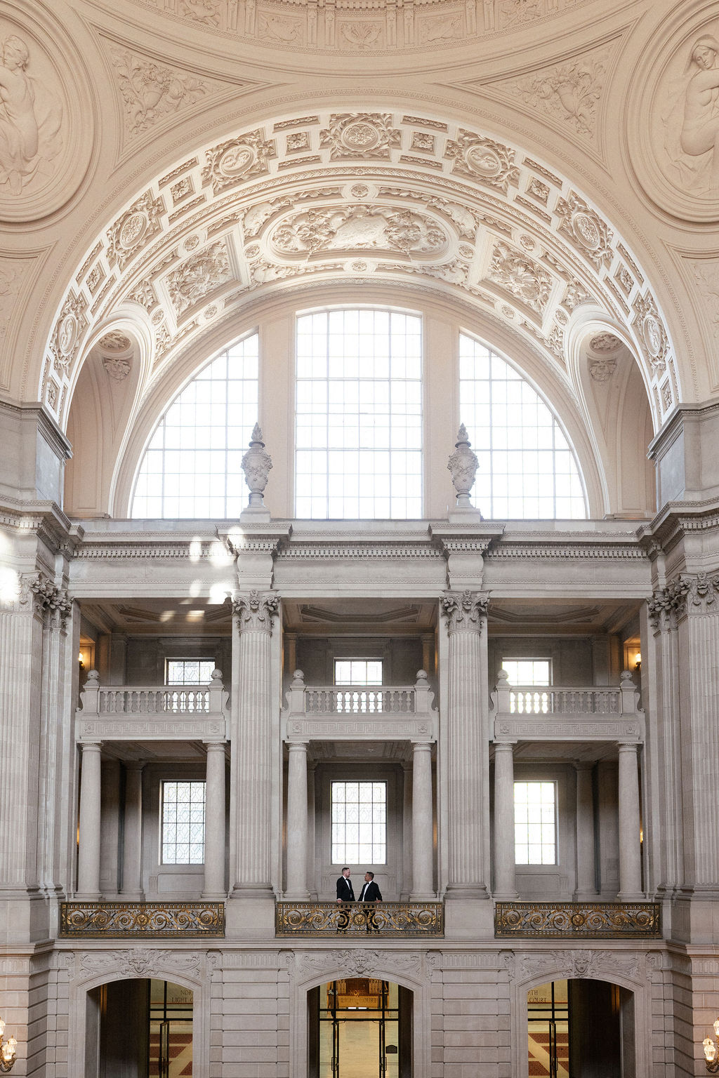 Interior view of a grand building with ornate architectural details, large arched window, and two people standing on a balcony.
