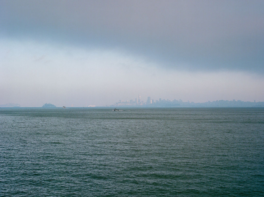 A calm body of water under a cloudy sky with a faint city skyline visible in the distance.