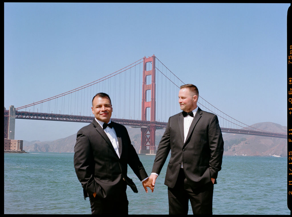 Two men in tuxedos hold hands and smile in front of the Golden Gate Bridge on a clear day.