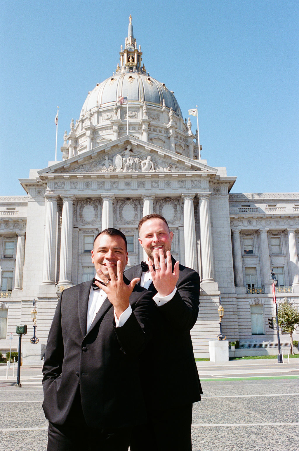 Two men in suits stand in front of a government building, smiling and showing wedding rings on their hands.
