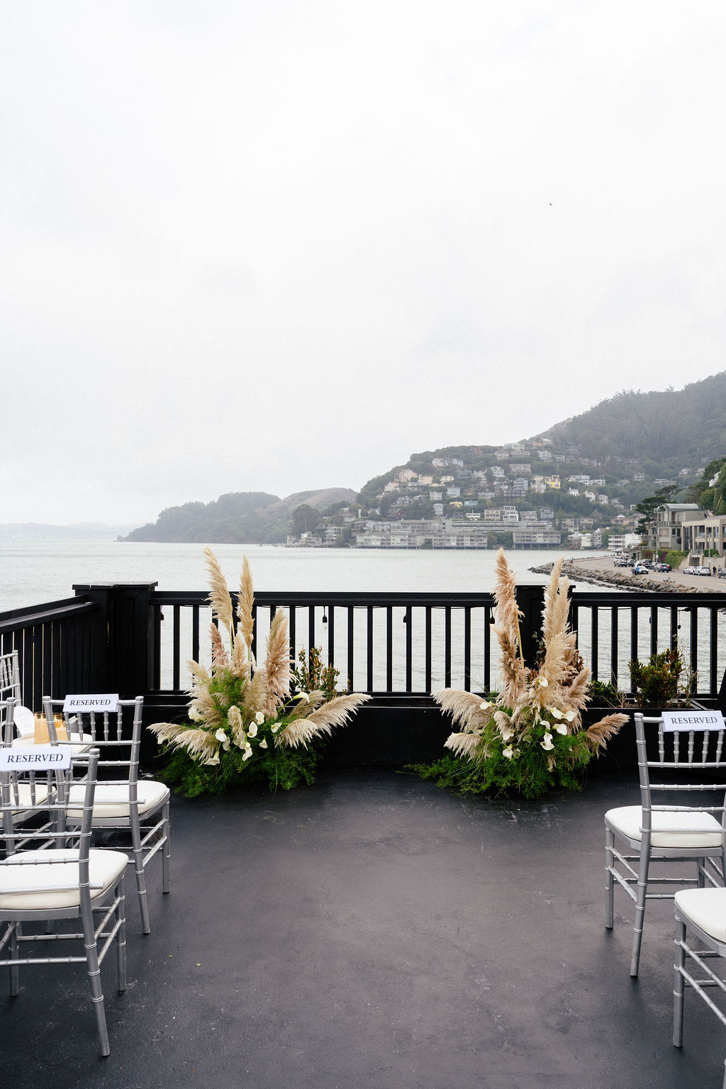 Outdoor deck with white chairs and pampas grass arrangements overlooks a body of water and hillsides with buildings in the distance on a cloudy day.