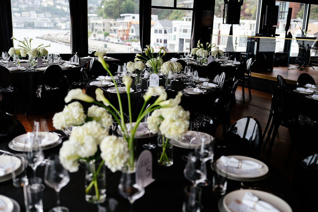 Round tables set for an event with white floral centerpieces, glassware, and plates, in a room with large windows overlooking buildings and water.