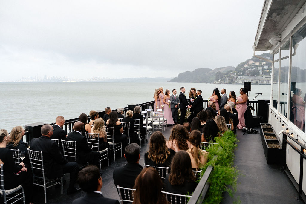 Outdoor wedding ceremony on a deck overlooking the water, with guests seated facing a couple and their wedding party under a cloudy sky.