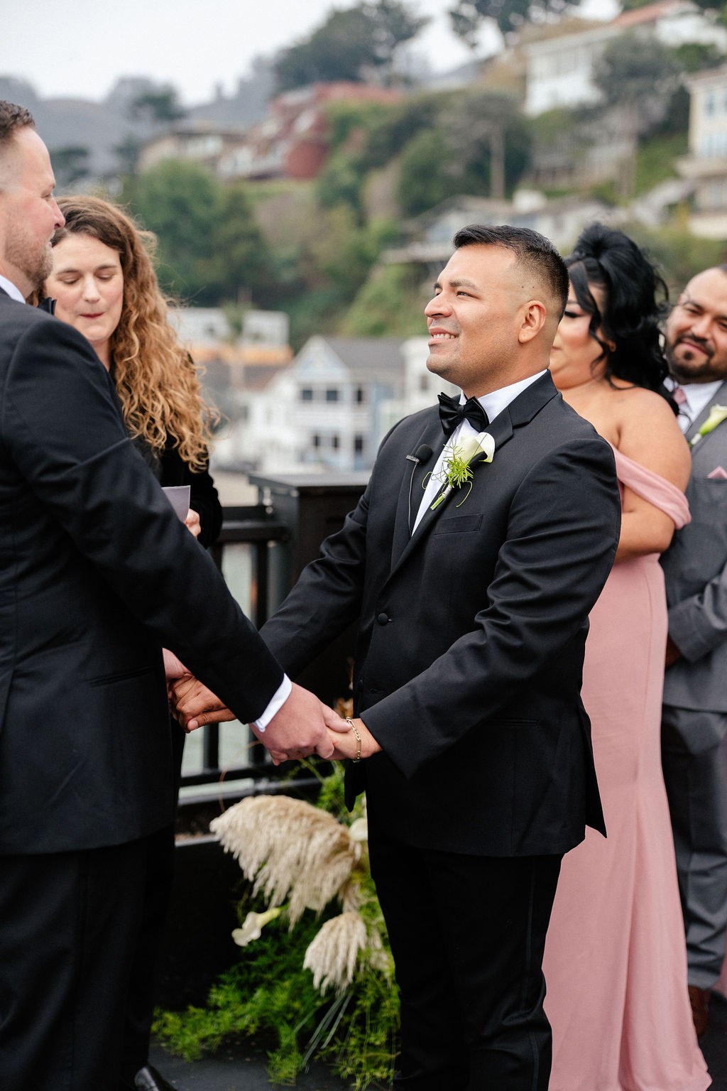 Two grooms in suits hold hands during an outdoor wedding ceremony, with bridesmaids and groomsmen standing behind them.