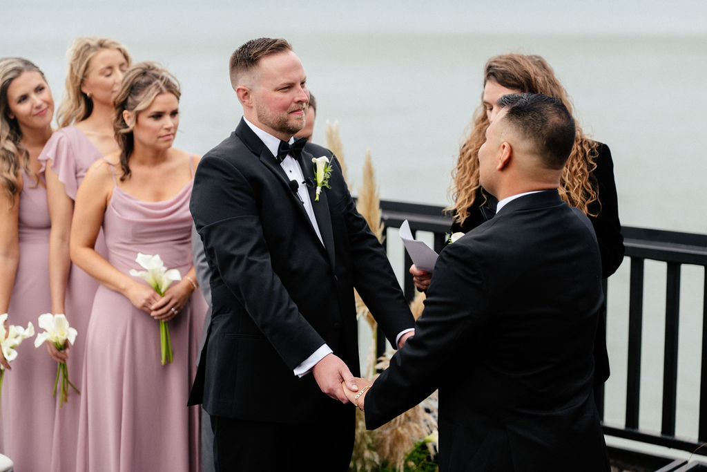 Two men in suits hold hands during an outdoor wedding ceremony by the water, with bridesmaids in mauve dresses standing beside them.