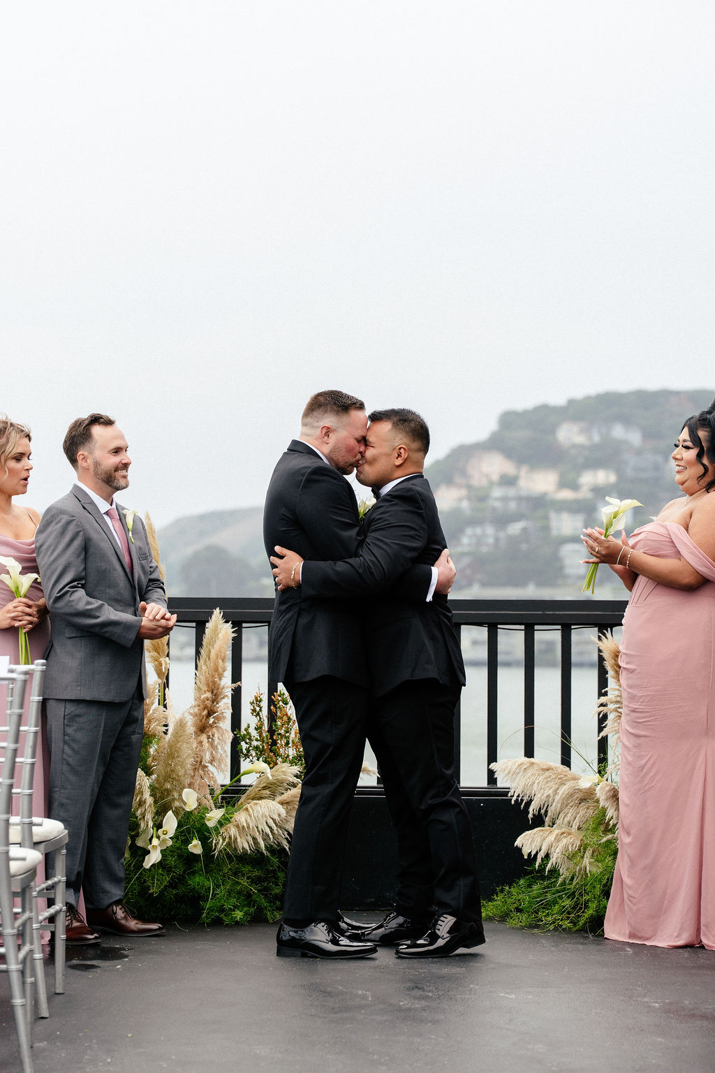 Two men in suits kiss at an outdoor wedding ceremony while their wedding party stands nearby; a cloudy landscape and water are visible in the background.
