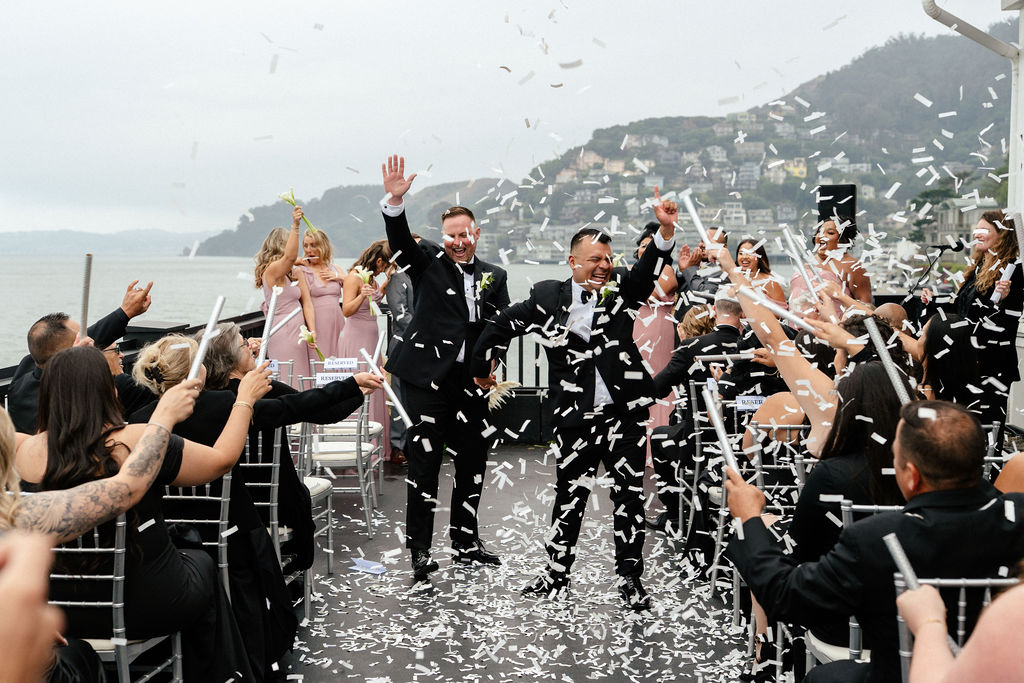 Two grooms in tuxedos walk down an outdoor aisle as guests celebrate with confetti; hills and water are visible in the background.