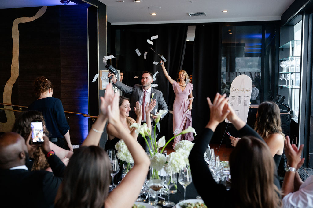 A couple enters a room celebrating as guests seated at decorated tables cheer and raise their hands; one guest takes a photo on their phone.