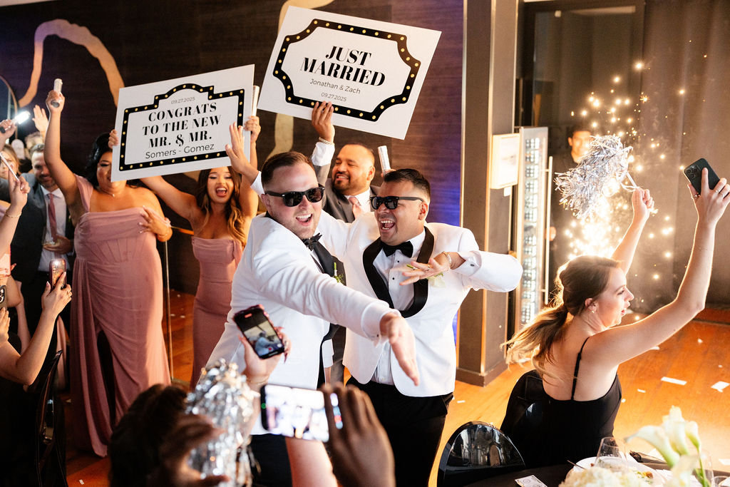 Two men in tuxedos and sunglasses celebrate at a wedding reception, holding signs that read "JUST MARRIED" and "CONGRATS TO THE NEW MR. &amp; MR.", surrounded by cheering guests.