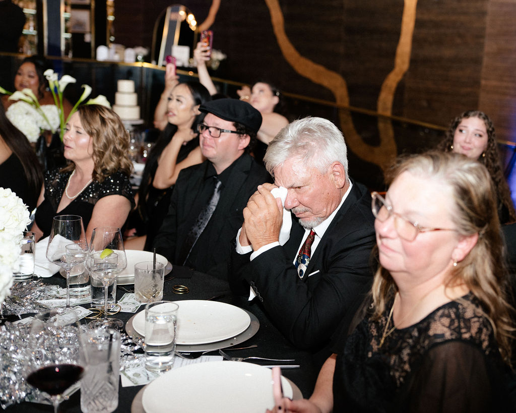 A group of people in formal attire sit at a decorated table; one man wipes his eyes with a tissue, while others watch something off camera.