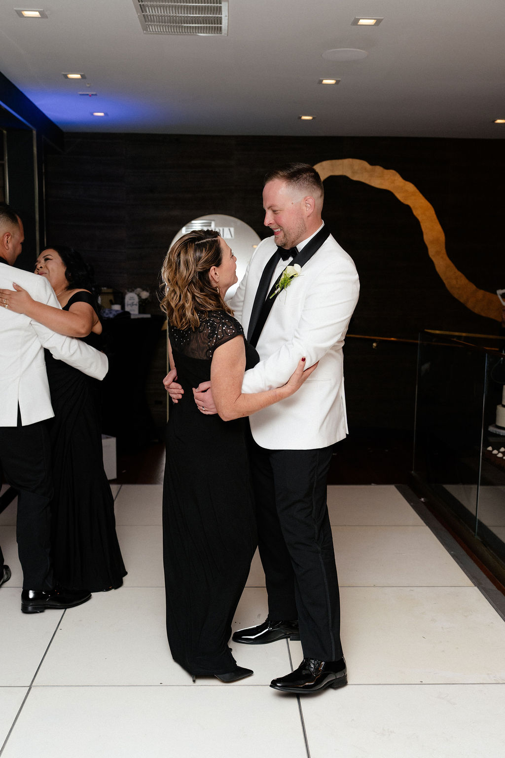 A man in a white tuxedo jacket and a woman in a black dress dance together at an indoor event, with another couple dancing in the background.