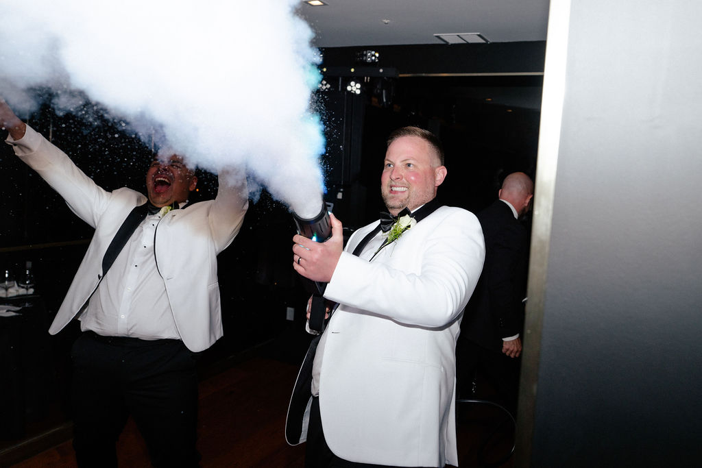 Two men in white tuxedos, one holding and firing a smoke machine, creating a large cloud indoors.