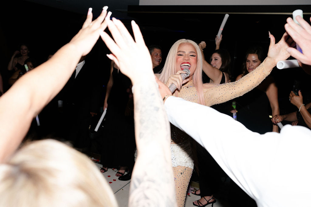 A performer with long pink hair sings into a microphone while surrounded by people reaching their hands toward her in a dark room.