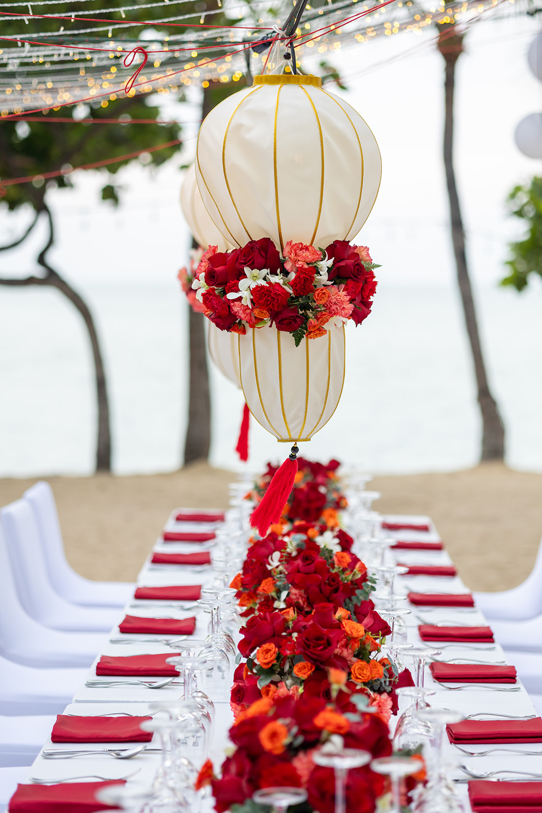 A long outdoor dining table with white chairs, red napkins, and a centerpiece of red and orange flowers under large white lanterns and string lights by the beach.