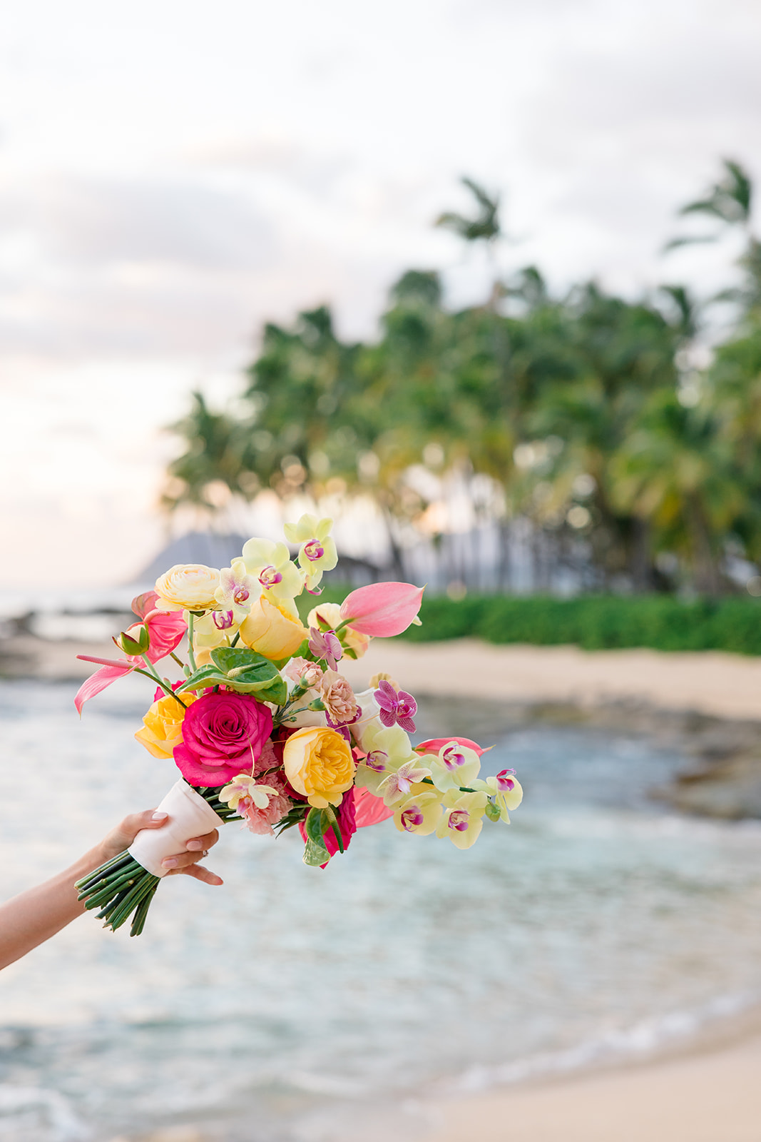 A hand holds a colorful bouquet of flowers by the ocean, with palm trees and a sandy beach in the background.