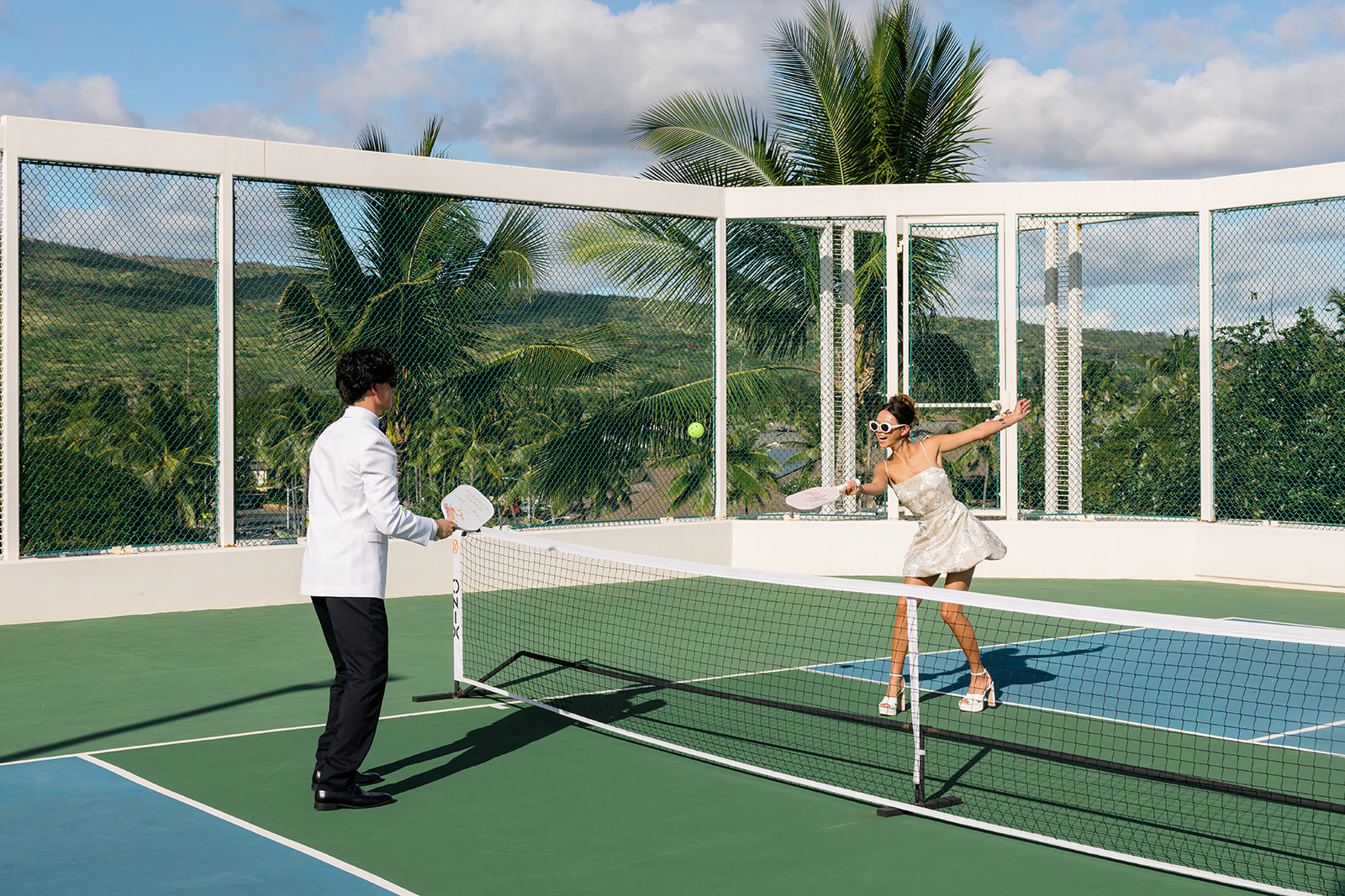Two people dressed formally play pickleball on an outdoor court surrounded by glass panels with a scenic landscape in the background.