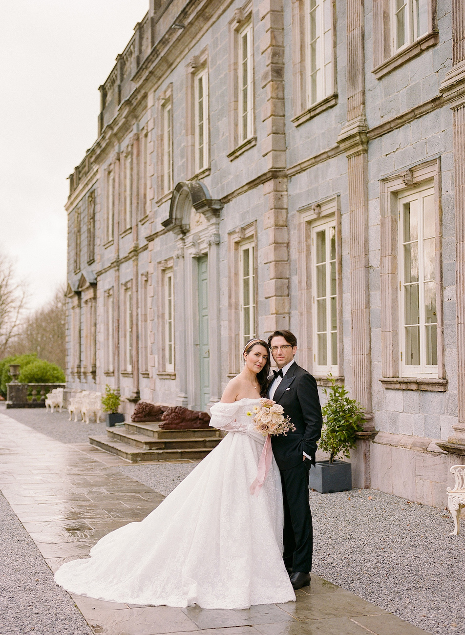 A bride and groom stand together in wedding attire outside a large, historic stone building with tall windows and overcast skies.