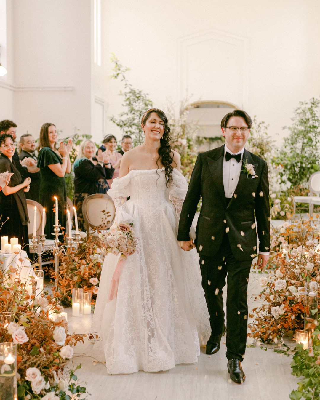 A bride in a white gown and a groom in a black tuxedo walk down the aisle, surrounded by guests, flowers, and candles in a decorated indoor venue.