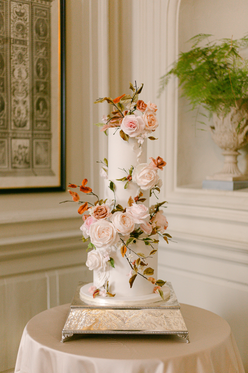 A two-tier white cake decorated with pink and orange flowers sits on a round table in an elegant room.